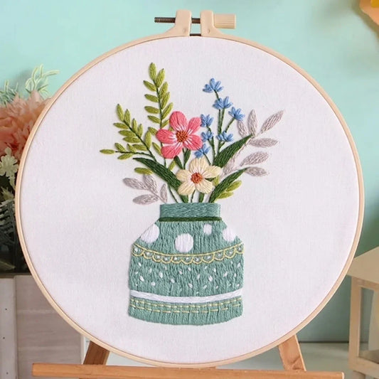 Hand embroidery of colorful flowers in a green vase on white fabric in a wooden hoop.