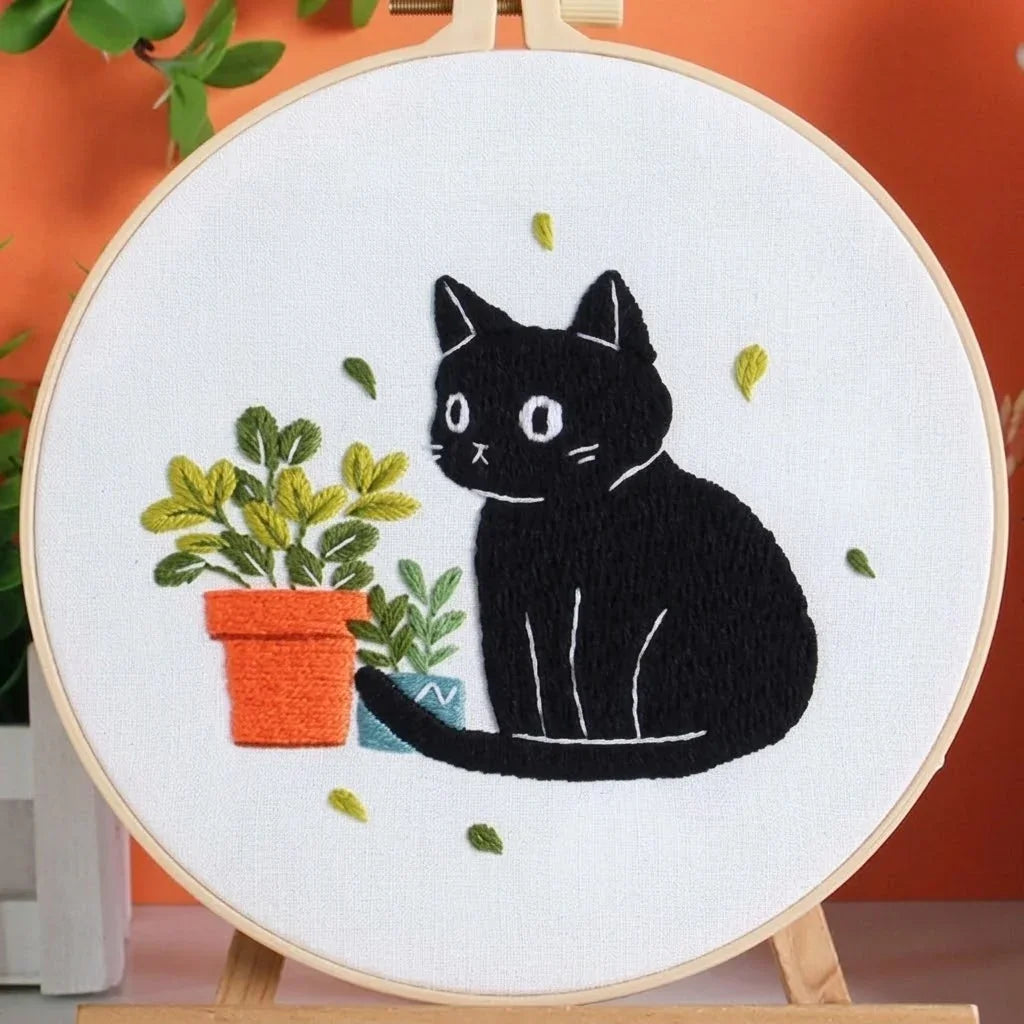 Embroidered hoop art featuring a black cat sitting next to potted green plants.