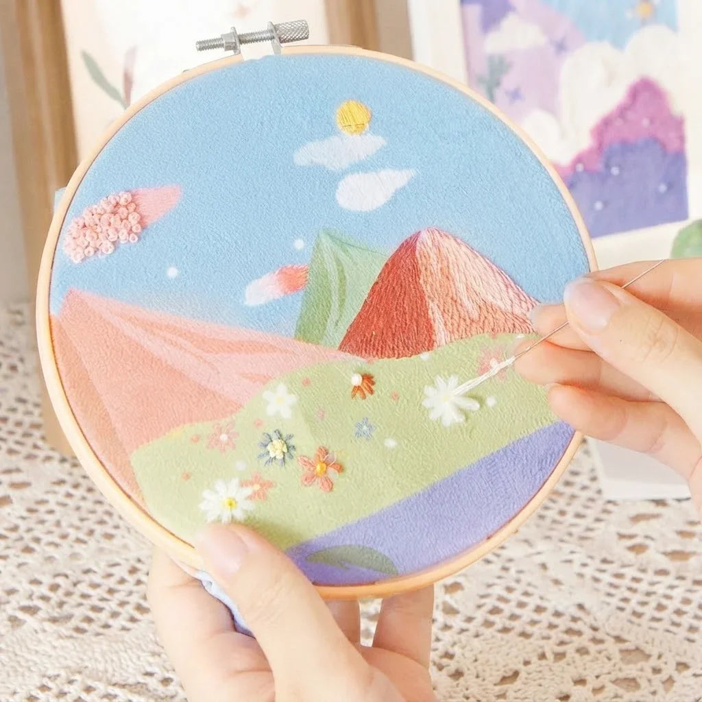 Hands stitching a landscape DIY embroidery painting kit with colorful mountains and flowers.
