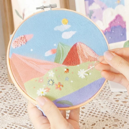 Hands stitching a landscape DIY embroidery painting kit with colorful mountains and flowers.