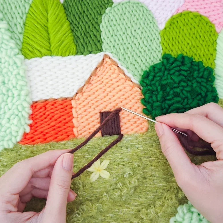 Hands using a needle to embroider a brown door on a Cabin in the Woods punch needle kit.