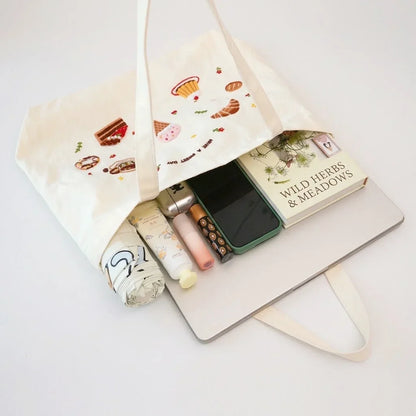 Cream tote bag with dessert embroidery containing a book, smartphone, and beauty products.