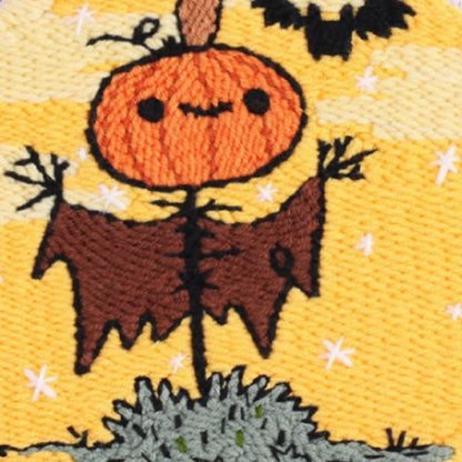 This punch needle design features a cheerful pumpkin-headed scarecrow under a golden Halloween sky, with a playful bat flying nearby. I