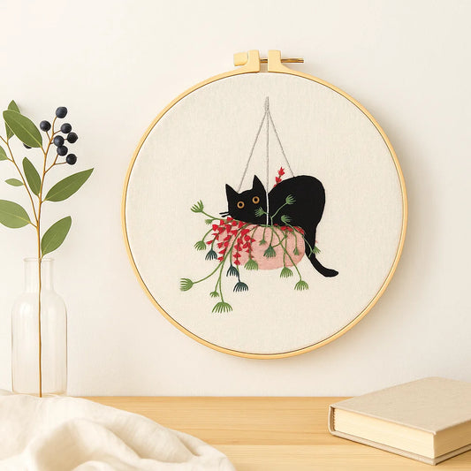 Embroidery hoop with a black cat design hanging from plants on a light background