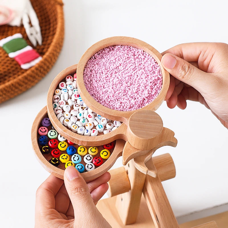 Wooden toy with compartments filled with colorful beads and pink sand, held by hands on a white background.