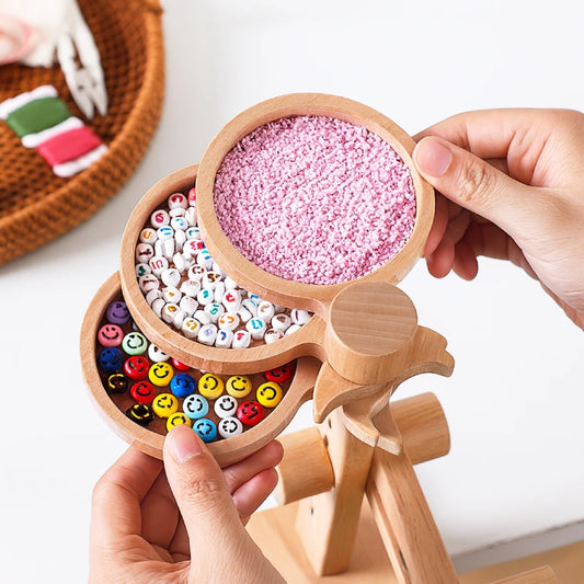 Wooden toy with compartments filled with colorful beads and pink sand, held by hands on a white background.