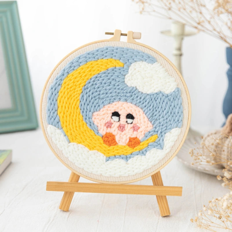 Handmade crochet artwork of a pink creature on a yellow crescent moon with a blue sky, displayed in a wooden hoop.