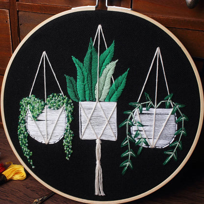 Embroidery hoop wall art kit with completed floral hanging planter design, featuring greenery and white embroidery on a black background.