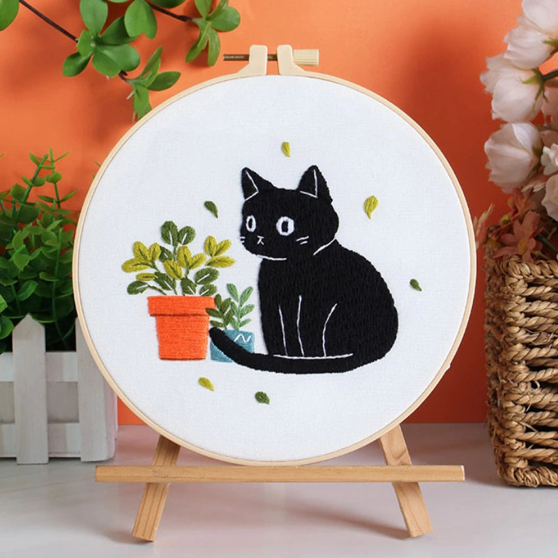 Embroidery hoop with a black cat design on a white background, placed on a wooden stand against an orange wall.