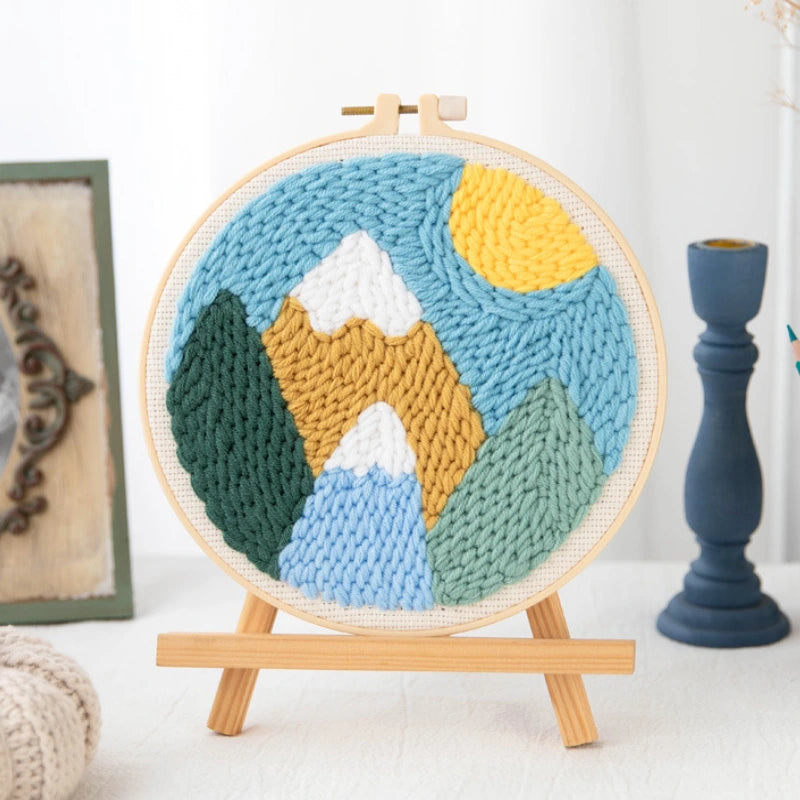 Knitted landscape scene in a wooden hoop on a white surface with a candle and framed picture in the background.