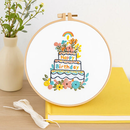Embroidery hoop with a colorful 'Happy Birthday' cake design on a white background, placed on a wooden surface with a yellow fabric and plant in the background.