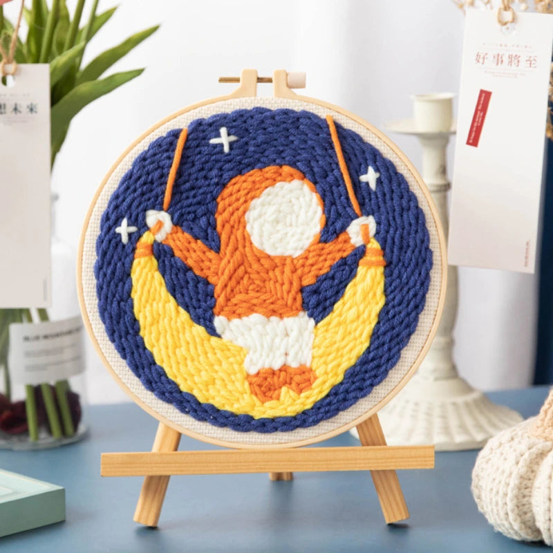 Embroidery hoop with a colorful design of a person on a moon, placed on a table.