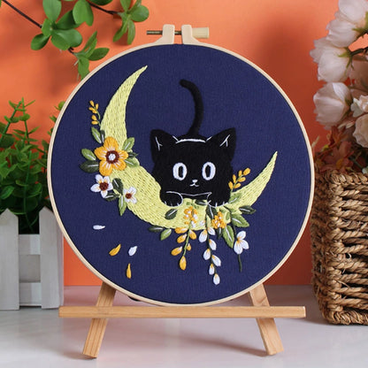 Embroidery hoop with a black cat on a crescent moon surrounded by flowers, placed on a wooden stand against an orange wall.