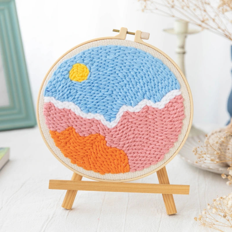 Woven artwork of a beach scene with a blue sky, pink sand, and orange water on a wooden stand.