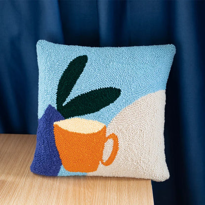 A punch needle pillow showing a bright orange cup beside green leaves under a clear blue sky.