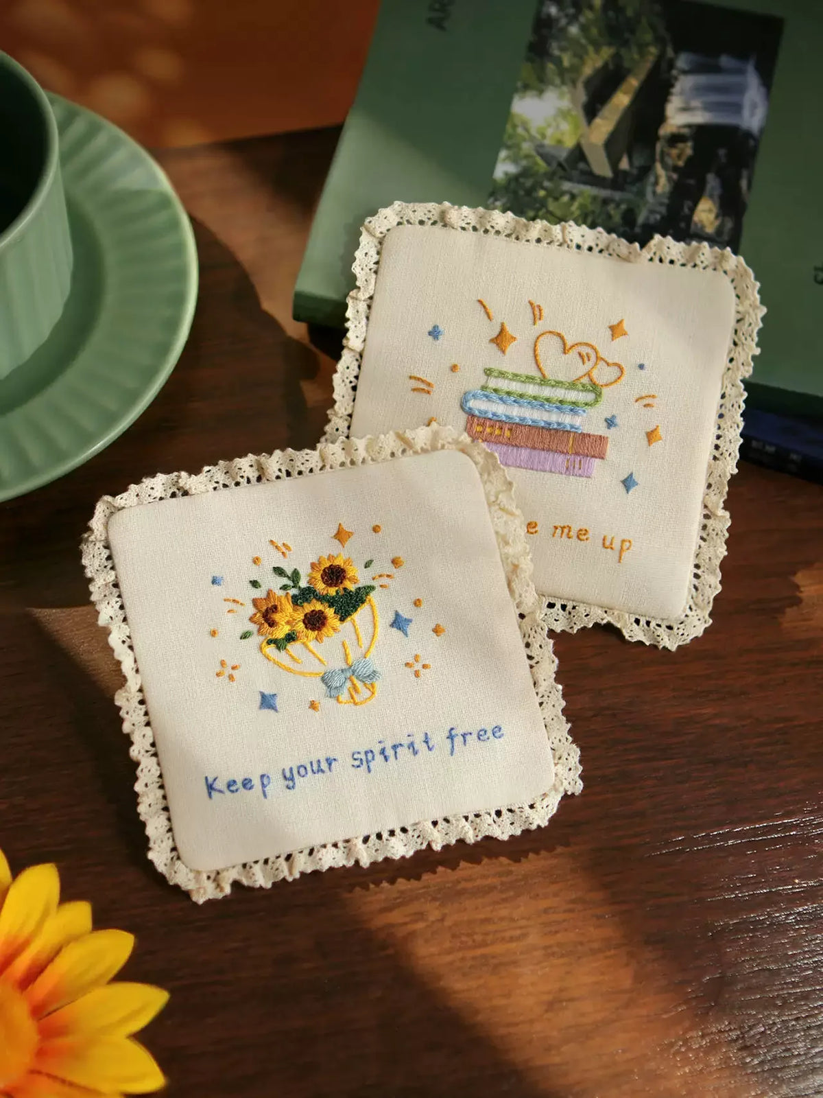 Two handmade embroidered coasters with floral and book designs and lace edges on a wooden table.