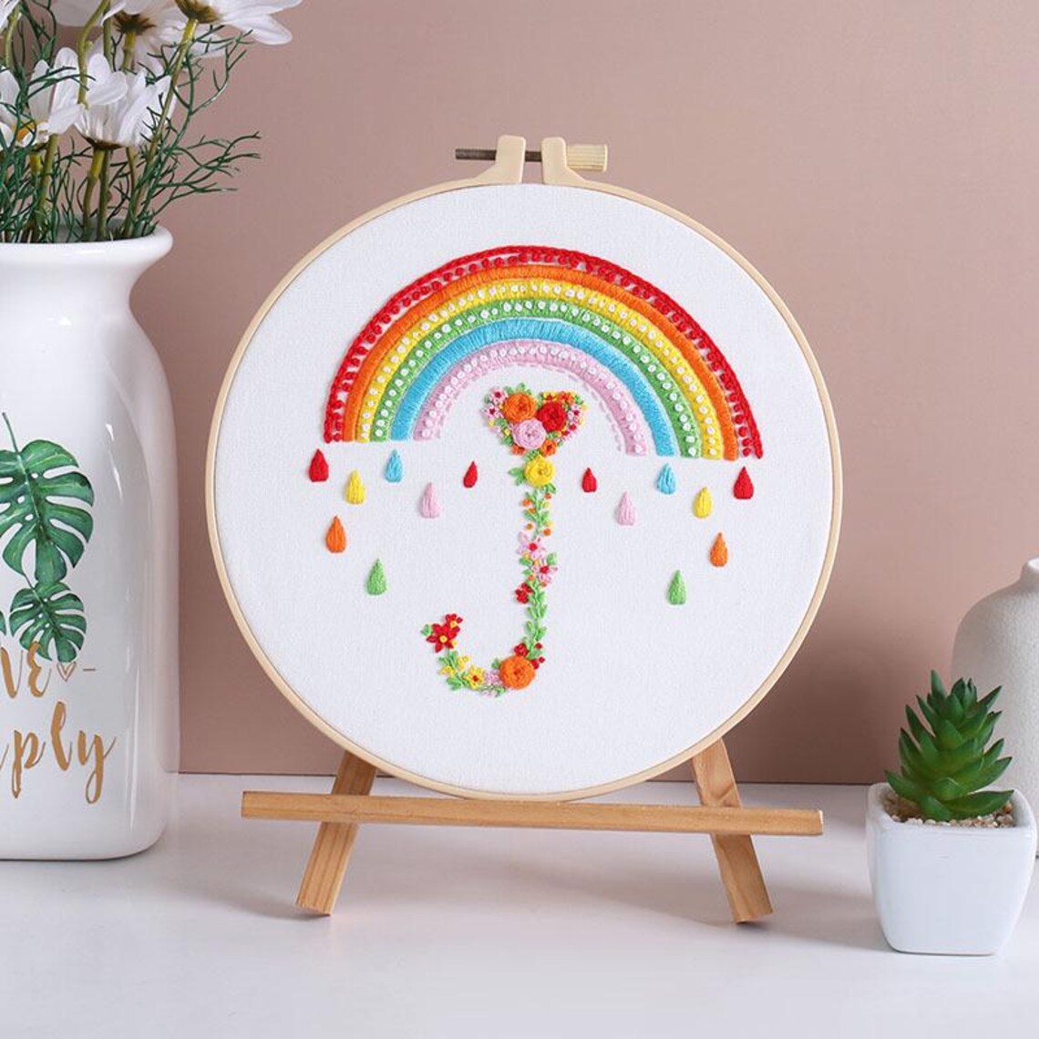 Embroidery hoop with a colorful rainbow and floral design on a wooden stand against a beige wall.
