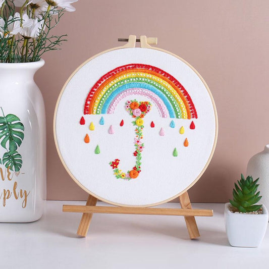 Embroidery hoop with a colorful rainbow and floral design on a wooden stand against a beige wall.