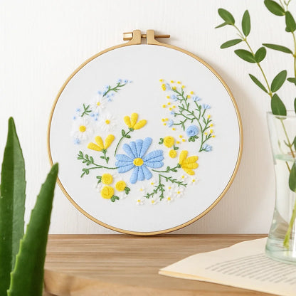 Embroidery hoop with floral design on a white background, placed on a wooden surface with plants around.