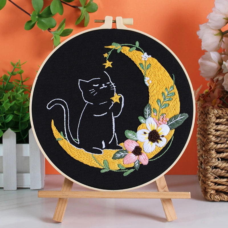 Embroidery hoop with a cat design on a crescent moon surrounded by flowers against an orange background.