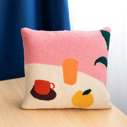 A punch needle pillow with a pink and beige background, featuring a cup, fruit, and glass in warm, cheerful tones.
