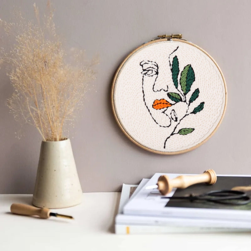 A minimalist face entwined with green leaves and orange lips, whispering softly to nature.The Punch Needle  on the wall.