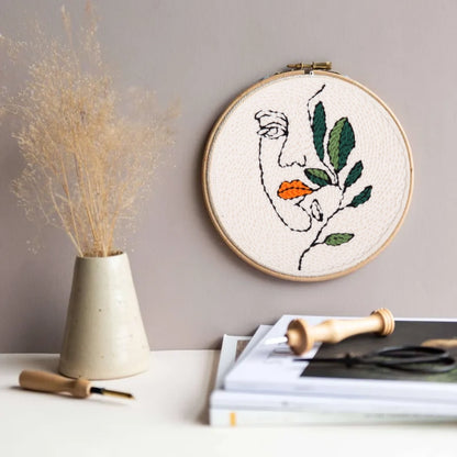 A minimalist face entwined with green leaves and orange lips, whispering softly to nature.The Punch Needle  on the wall.