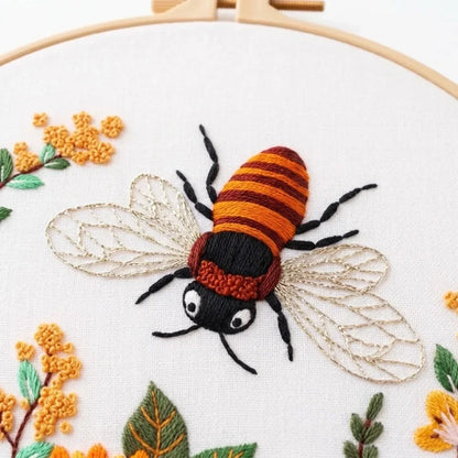 Detailed embroidery of a honey bee with orange stripes and gold wings on a white fabric hoop.