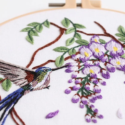 Embroidery of a hummingbird feeding on purple flowers on a branch.