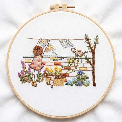 Hand embroidery of a girl hanging laundry with cats and flowers in a garden setting.