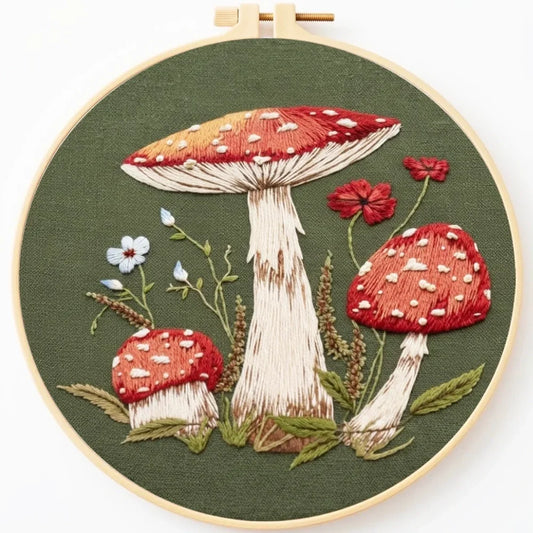 Hand embroidery of red amanita mushrooms and wildflowers on green fabric in a wooden hoop.